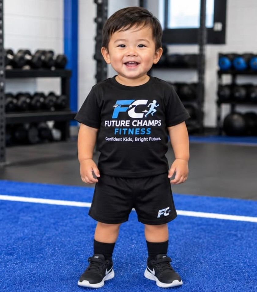 A smiling toddler in a Future Champs Fitness shirt standing on blue turf